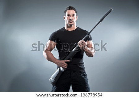 Action hero muscled man holding a rifle. Wearing black t-shirt and pants. Studio shot against grey.