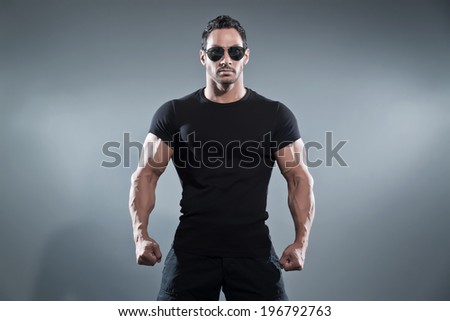 Combat muscled action hero man wearing black t-shirt with pants and sunglasses. Studio shot against grey.