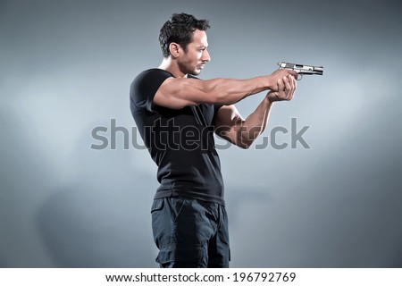 Action hero muscled man shooting with gun. Wearing black t-shirt and pants. Studio shot against grey.