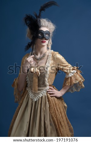 Retro baroque fashion woman wearing gold dress. Holding a black mask. Studio shot against blue.