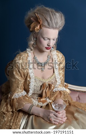 Retro baroque fashion woman wearing gold dress. Holding wine glass. Sitting on vintage couch. Studio shot against blue.