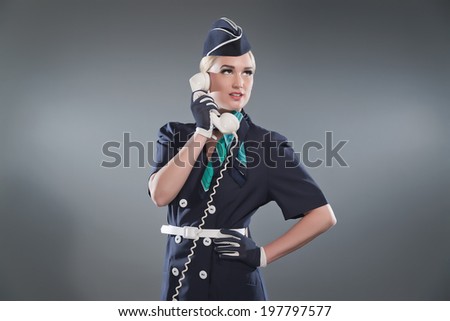 Calling retro blonde stewardess wearing blue suit. Holding white vintage phone. Studio shot against grey.