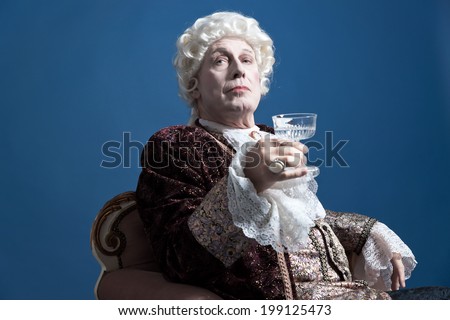 Retro baroque man with white wig holding a wine glass sitting on antique couch. Studio shot against blue.