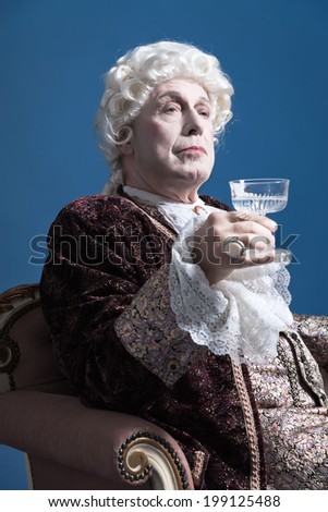 Retro baroque man with white wig holding a wine glass sitting on antique couch. Studio shot against blue.