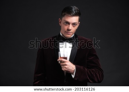 Well dressed good looking business man holding glass of whiskey. Wearing dark red dinner jacket and black bow tie.