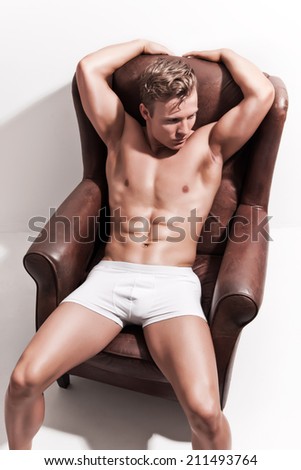 Male muscled underwear model wearing white shorts. Blonde hair. Sitting in leather chair against white wall.