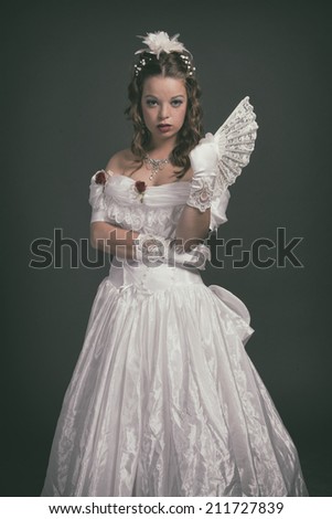 Victorian fashion woman wearing white dress. Holding fan. Studio shot against grey.