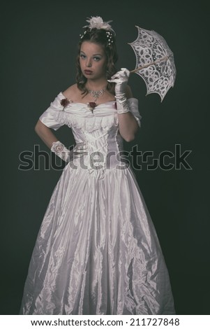 Victorian fashion woman wearing white dress. Holding parasol. Studio shot against grey.