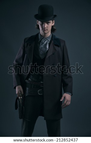 Western 1900 fashion man with brown hair and hat holding gun. Studio shot against grey.