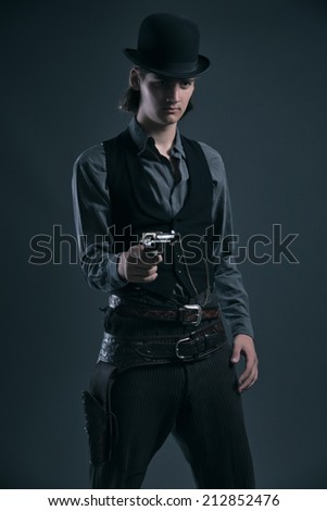 Western 1900 fashion man with brown hair and hat shooting with gun. Studio shot against grey.