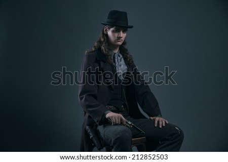 Western 1900 fashion man with brown hair and hat sitting on chair holding gun. Studio shot against grey.