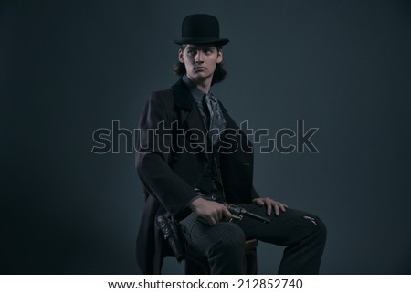 Western 1900 fashion man with brown hair and hat sitting on chair holding gun. Studio shot against grey.