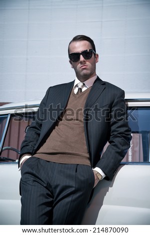 Retro 60s fashion business man wearing grey suit and black sunglasses standing against classic car.