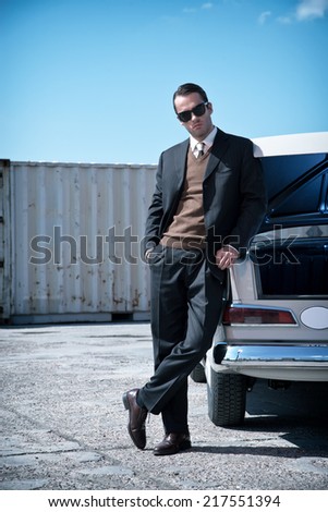 Retro fifties mafia fashion man standing next to open trunk of vintage car.