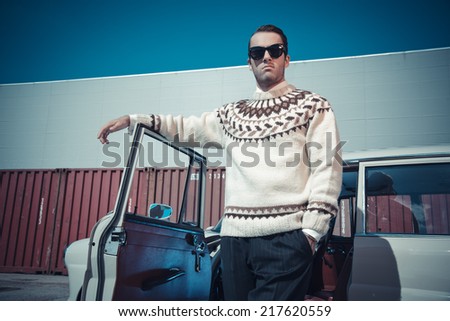 Retro fifties fashion man with woolen sweater and sunglasses standing against vintage car.