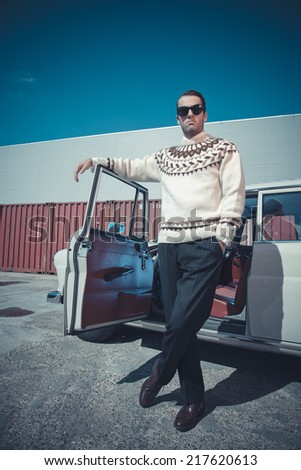 Retro fifties fashion man with woolen sweater and sunglasses standing against vintage car.