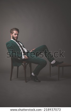 Business fashion man wearing green suit with white shirt black and tie. Sitting on wooden chair. Studio shot against grey.
