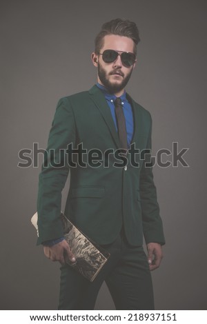 Business fashion man wearing green suit with sunglasses and blue shirt and black tie. Holding a magazine. Studio shot against grey.