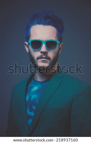 Man with blue hair and brown beard wearing green suit. Wearing blue sunglasses. Male fashion. Studio shot against dark background.