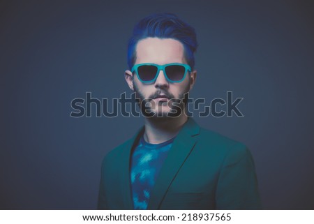 Man with blue hair and brown beard wearing green suit. Wearing blue sunglasses. Male fashion. Studio shot against dark background.