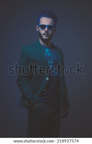 Man with blue hair and brown beard wearing green suit. Wearing blue sunglasses. Male fashion. Studio shot against dark background.