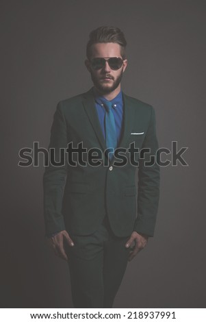 Business fashion man wearing green suit with sunglasses and blue shirt and tie. Studio shot against grey.