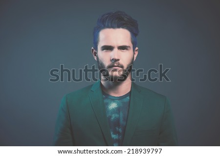Man with blue hair and brown beard wearing green suit. Male fashion. Studio shot against dark background.
