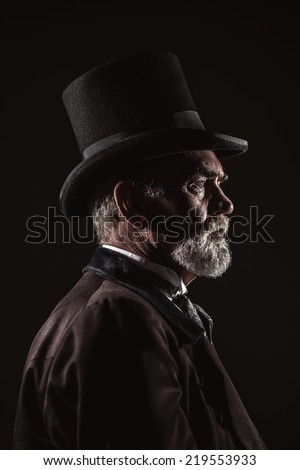 Vintage victorian man with black hat and gray hair and beard. Studio shot against dark background.