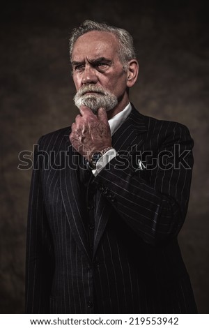 Characteristic senior business man with gray hair and beard wearing blue striped suit and tie. Against brown wall.