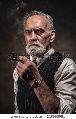 Cigar smoking characteristic senior business man with gray hair and beard wearing blue striped gilet and tie. Against brown wall.
