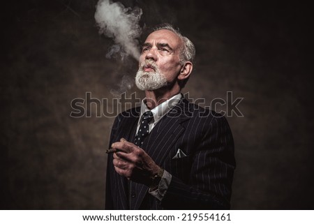 Cigar smoking characteristic senior business man with gray hair and beard wearing blue striped suit and tie. Against brown wall.
