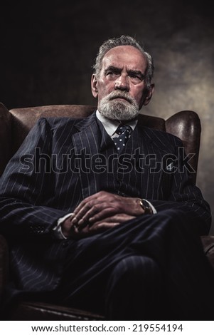 In chair sitting characteristic senior business man. Gray hair and beard wearing blue striped suit and tie. Against brown wall.