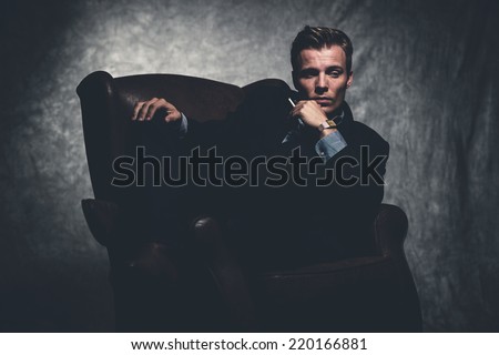 Cigarette smoking retro fifties cool business fashion man wearing black suit. Sitting in leather chair. Gray wall.