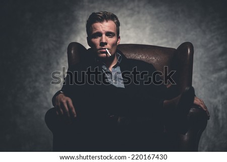 Cigarette smoking retro fifties cool business fashion man wearing black suit. Sitting in leather chair. Gray wall.
