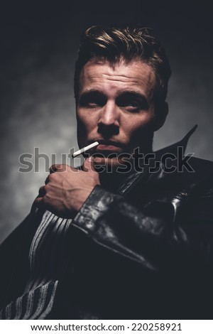 Cigarette smoking retro fifties cool rebellion fashion man wearing striped woolen sweater and black leather jacket. Gray wall.