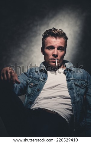 Cigarette smoking retro fifties cool fashion man wearing white shirt and jeans jacket. Gray wall.