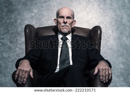 In leather chair sitting senior businessman with gray beard wearing dark suit and tie. Against grey wall.