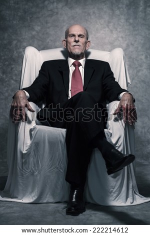 Bad businessman with bloody hands sitting in white chair. Gray beard wearing dark suit and red tie. Against grey wall.