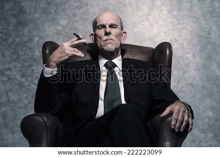 Cigar smoking senior businessman with gray beard wearing dark suit and tie. Sitting in leather chair. Against grey wall.