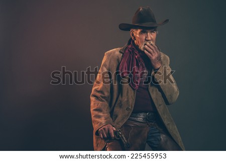 Old rough western cowboy with gray beard and brown hat smoking a cigarette. Low key studio shot.
