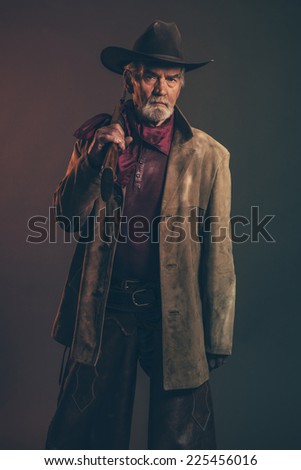 Old rough western cowboy with gray beard and brown hat holding rifle. Low key studio shot.