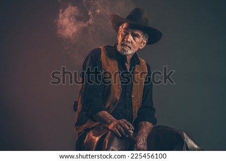 Cigarette smoking old rough western cowboy with gray beard and brown hat. Low key studio shot.