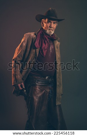 Old rough western cowboy with gray beard and brown hat pulling revolver. Low key studio shot.
