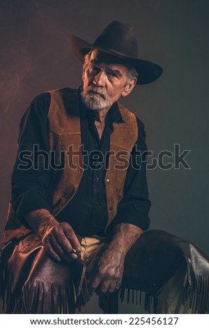 Cigarette smoking old rough western cowboy with gray beard and brown hat. Low key studio shot.