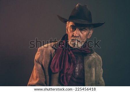 Old rough western cowboy with gray beard and brown hat smoking a cigarette. Low key studio shot.