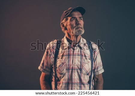 Characteristic senior man with gray hair and beard wearing blue cap with braces and jeans. Low key studio shot.
