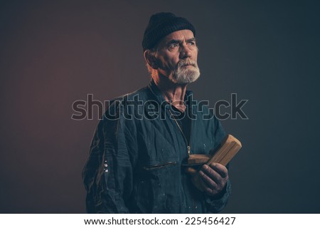Senior carpenter with gray hair and beard holding old plane wearing black hat with green overalls. Low key studio shot.