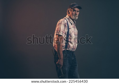 Senior male carpenter with gray hair and beard holding a hammer wearing blue cap with braces and jeans. Low key studio shot.
