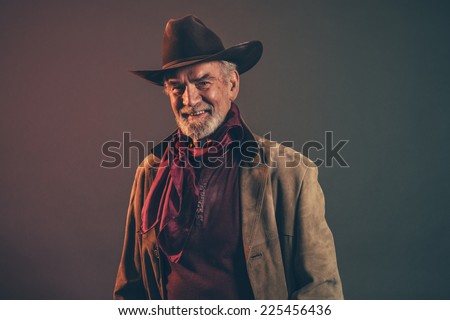 Smiling old rough western cowboy with gray beard and brown hat. Low key studio shot.