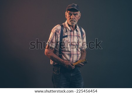 Senior carpenter with gray hair and beard holding yellow measuring rod wearing blue cap with braces and jeans. Low key studio shot.